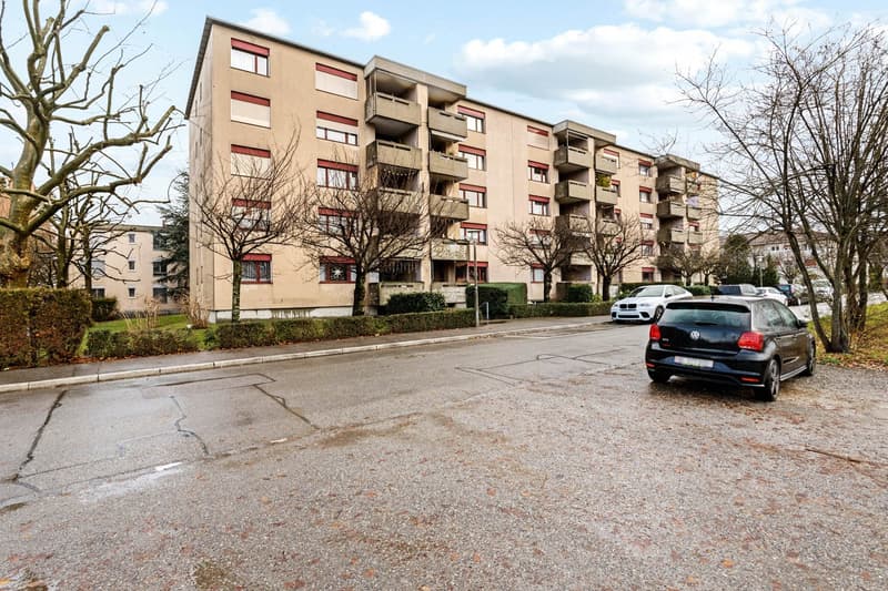 multi-story residential building, balconies, concrete exterior, parked cars, bare trees, street lights, bushes