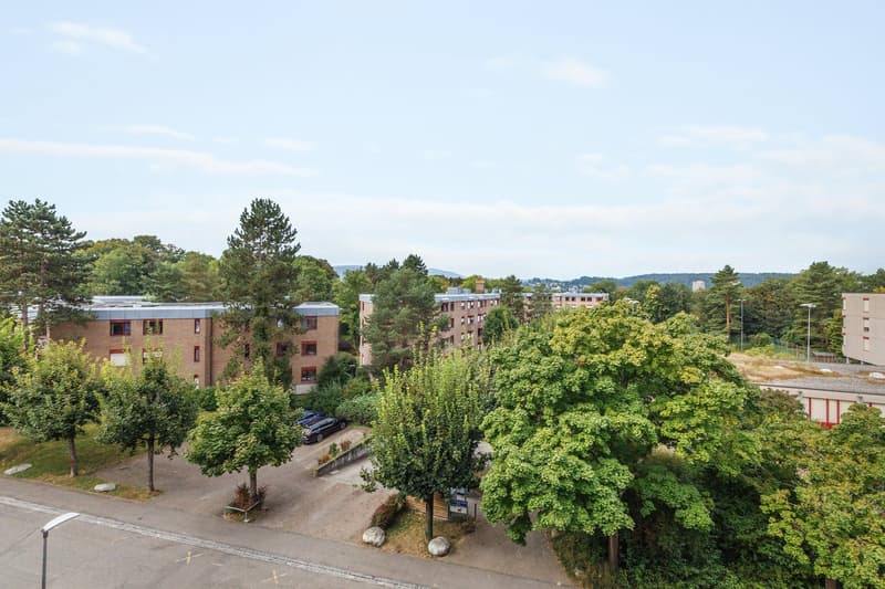 Three story residential building with parking lot in front of building. Trees in front of parking lot and around the building. A road visible on the left side of the image.