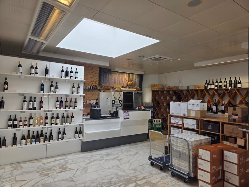 Interior of a wine shop. Counter with wine bottles on display shelves, wine racks, wine storage boxes on the floor, and a small cart.