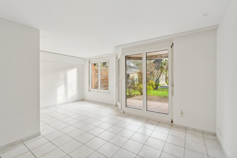 An empty room with white walls and white tiled flooring. There are two glass doors leading to a patio with a grassy area and trees. The room has a white ceiling and a power outlet on the wall.