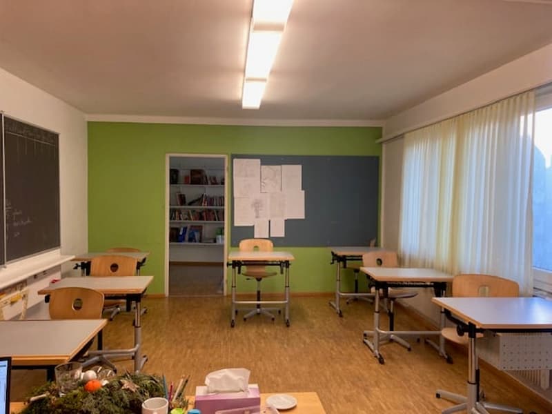 an empty classroom with wooden desks and chairs, green and white walls, bookshelves, and a projector screen