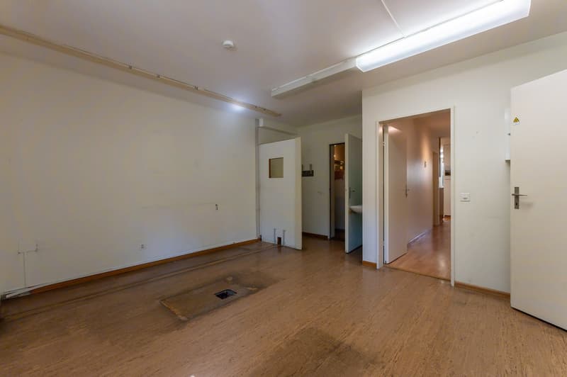 empty room, wooden floor, white walls, two doors, one leading to another room, one leading to a hallway, ceiling lights