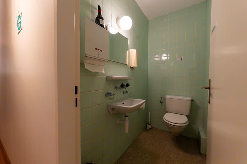 Small bathroom with green tiles, white sink, white toilet, and a mirror with a tissue dispenser on top