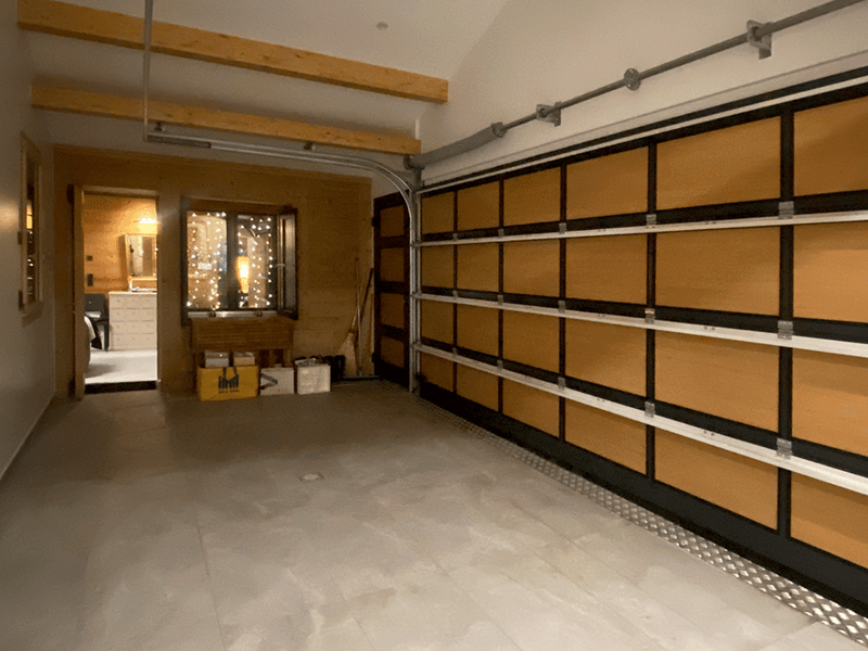 The image shows a spacious garage with wooden beams on the ceiling, a sliding garage door, and shelving units along the walls for storage.