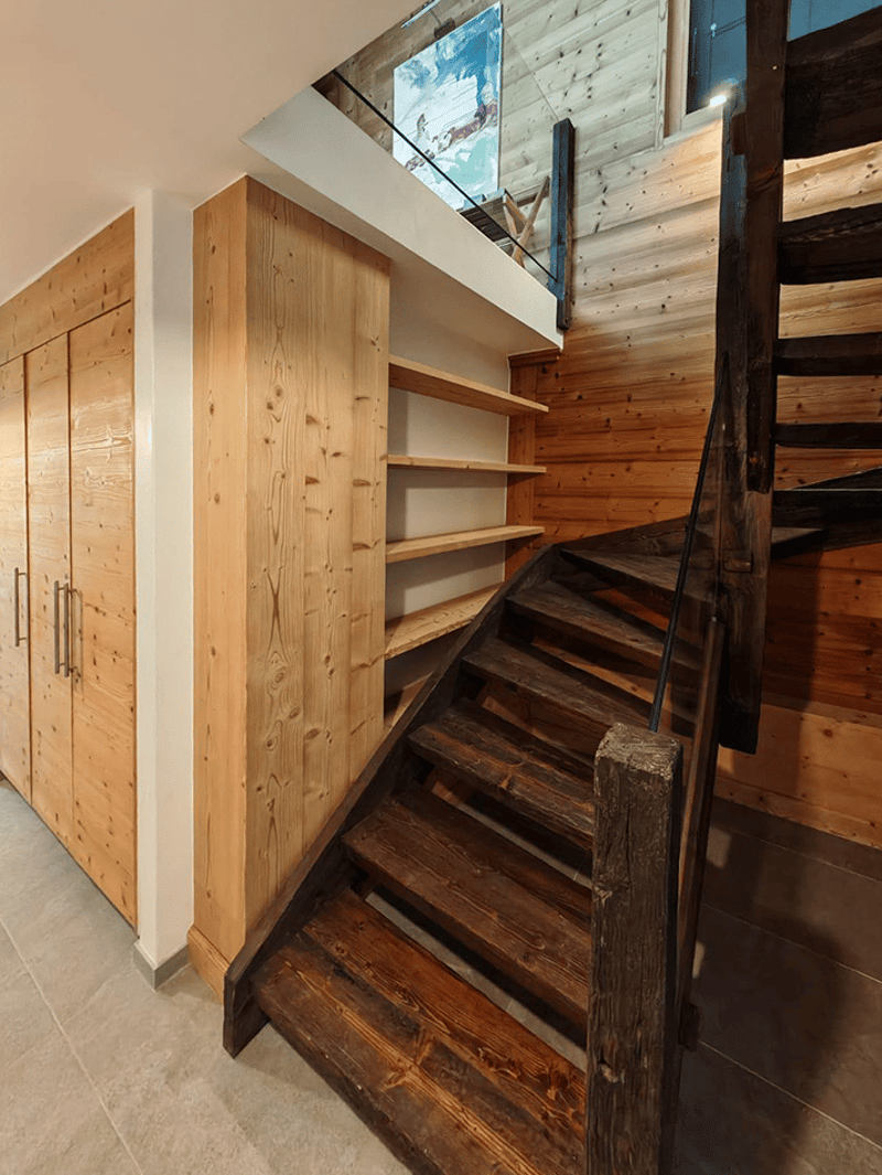 The image shows a rustic, wooden staircase leading up to a loft or upper level. The staircase is made of dark, weathered wood and has a sturdy, wooden railing. The walls and ceiling are also made of wood, giving the space a cozy, cabin-like feel. There is