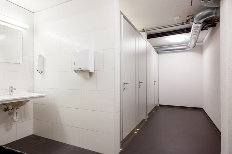 Tiled bathroom with white walls, a sink, and multiple stalls or cubicles with doors.