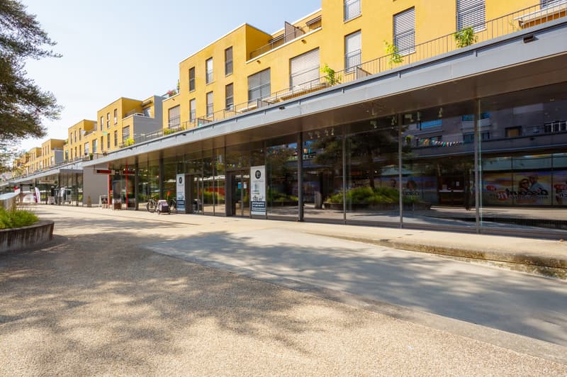 Multi-story commercial building with yellow exterior, glass storefronts, and balconies on the upper floors. The building has a modern, urban design and is situated in a pedestrian-friendly area with a paved walkway in front.