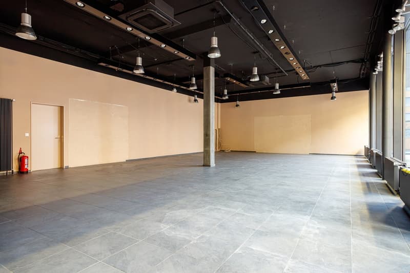 Large open commercial space with concrete flooring, exposed ceiling beams and ductwork, and track lighting. The space appears to be a studio or showroom with a fire extinguisher visible in the corner.