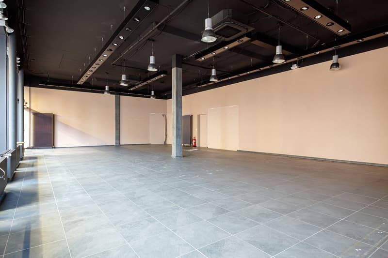 Large open commercial space with high ceilings, exposed beams, and track lighting. The space has a concrete floor with gray tiles and white walls. There is a fire extinguisher visible in the corner.