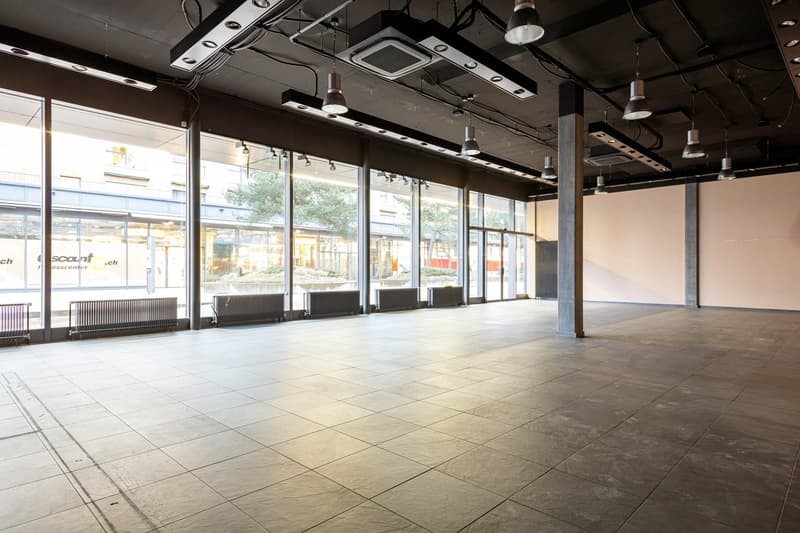 Large open commercial space with high ceilings, concrete floors, and large windows overlooking the exterior. The space has a modern, industrial design with exposed ductwork and lighting fixtures.