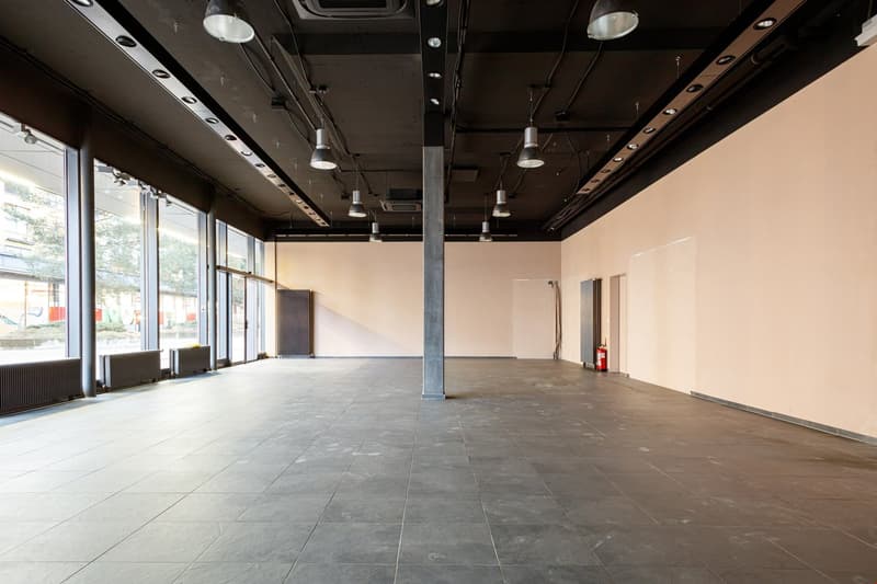 Large open commercial space with concrete flooring, exposed ceiling with lighting fixtures, and large windows along one wall.