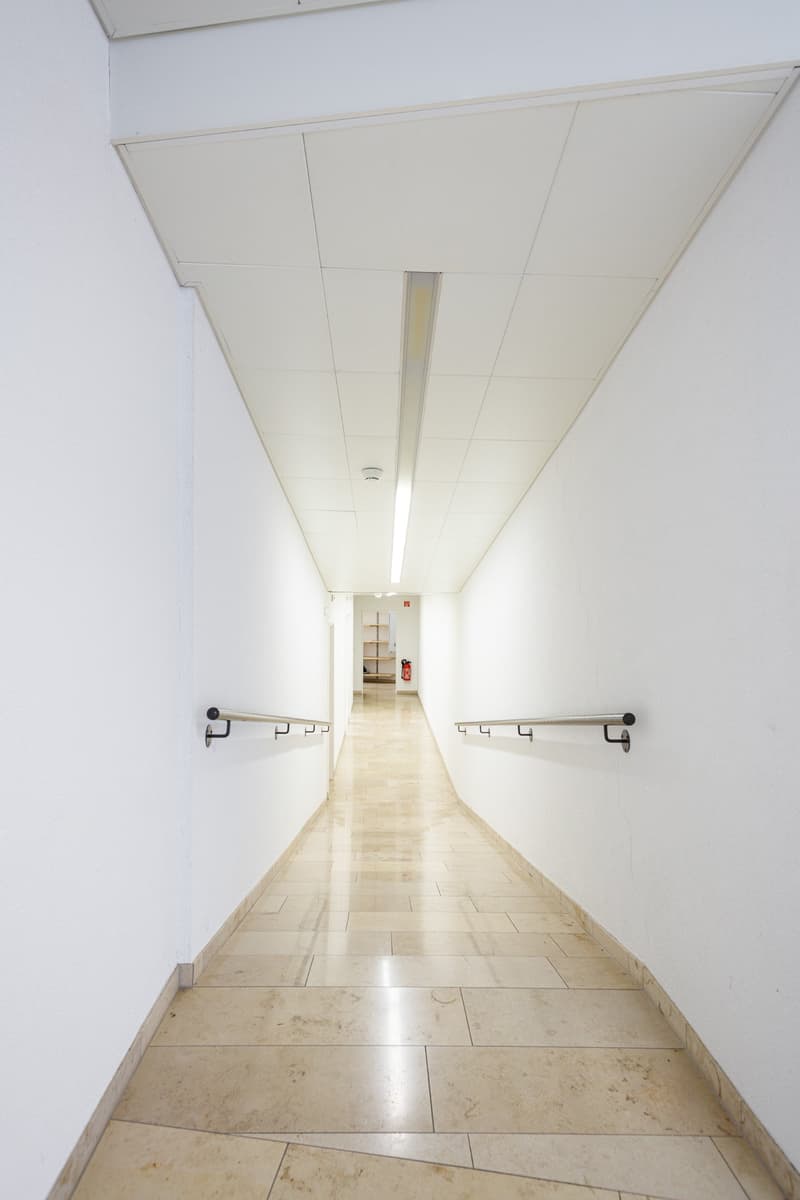 Empty hallway with white walls and tiled flooring, metal handrails, fire extinguisher, and ceiling lighting.