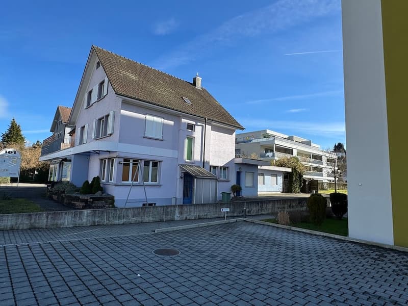 House with brown roof, white wall, balcony on second floor, multiple windows, front yard with a paved area
