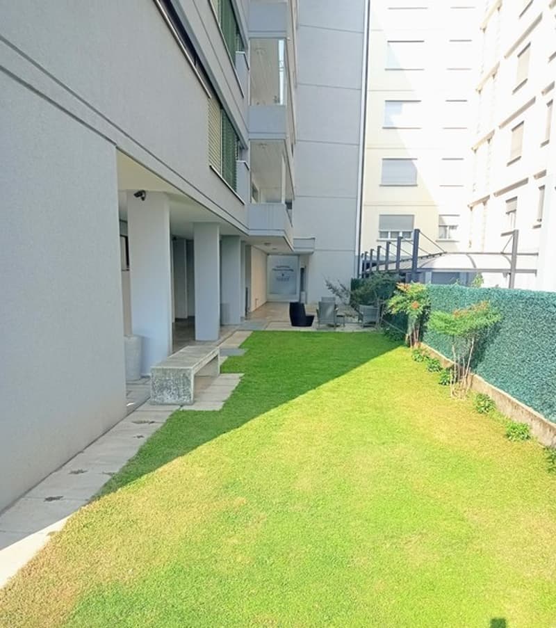 Multi-story apartment building with a well-maintained grassy area and landscaping in the foreground. The building has a modern, glass-and-concrete design with balconies visible on some floors.