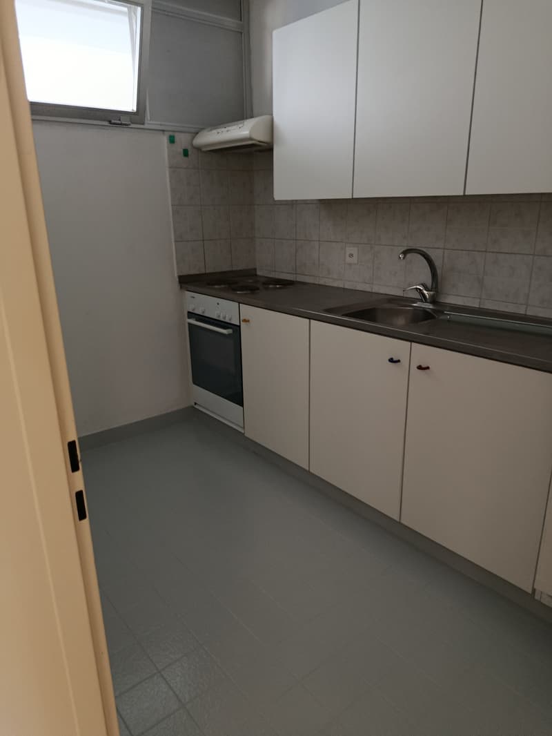 A kitchen with gray tile flooring, white walls, white ceiling, white cabinets, an oven, a sink, a faucet, and a power outlet. The kitchen also features a small window and a white appliance above the stove. The countertop is gray, and the floor is clean an