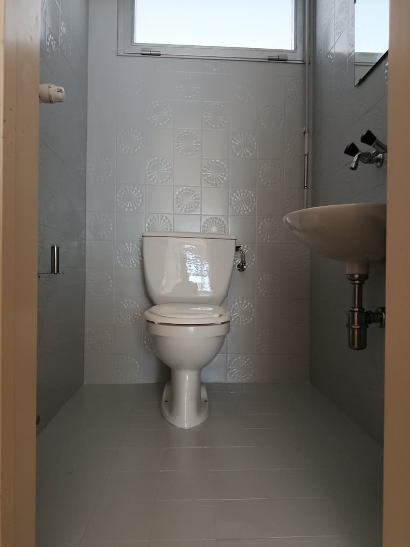 Small bathroom with a toilet and sink on the right side. The walls and floor are tiled with white decorative tiles. The sink has a silver faucet. There is a mirror on the wall above the sink. A small window provides natural light.