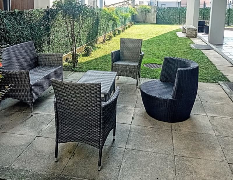 Patio with wicker furniture, including chairs and a table, overlooking a grassy lawn and garden area