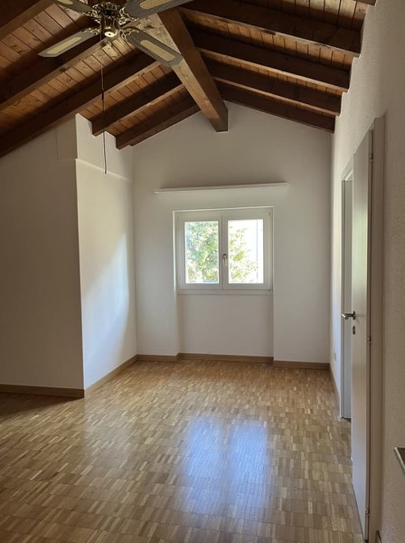 Spacious room with wooden ceiling beams, hardwood flooring, and a ceiling fan. The room has a large window providing natural light.
