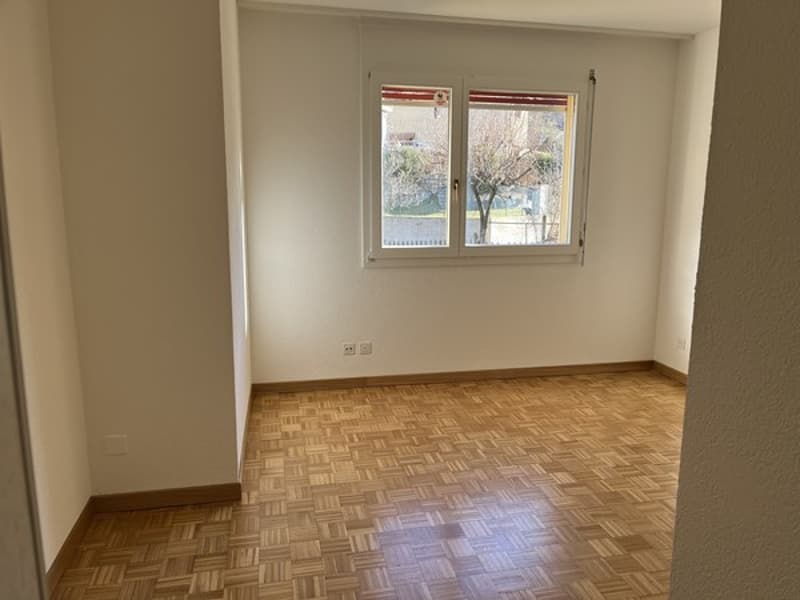 The image shows an empty room with white walls and a hardwood parquet floor. There are two windows that provide natural light to the space.