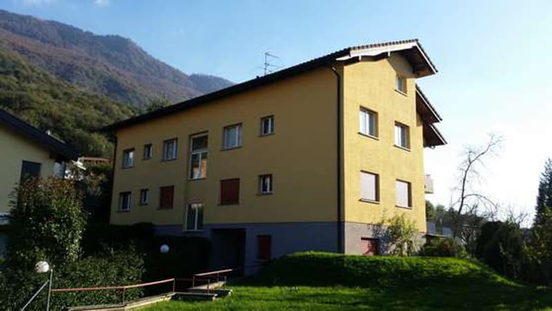 3-story yellow apartment building with balconies, surrounded by trees and greenery, with mountains in the background.