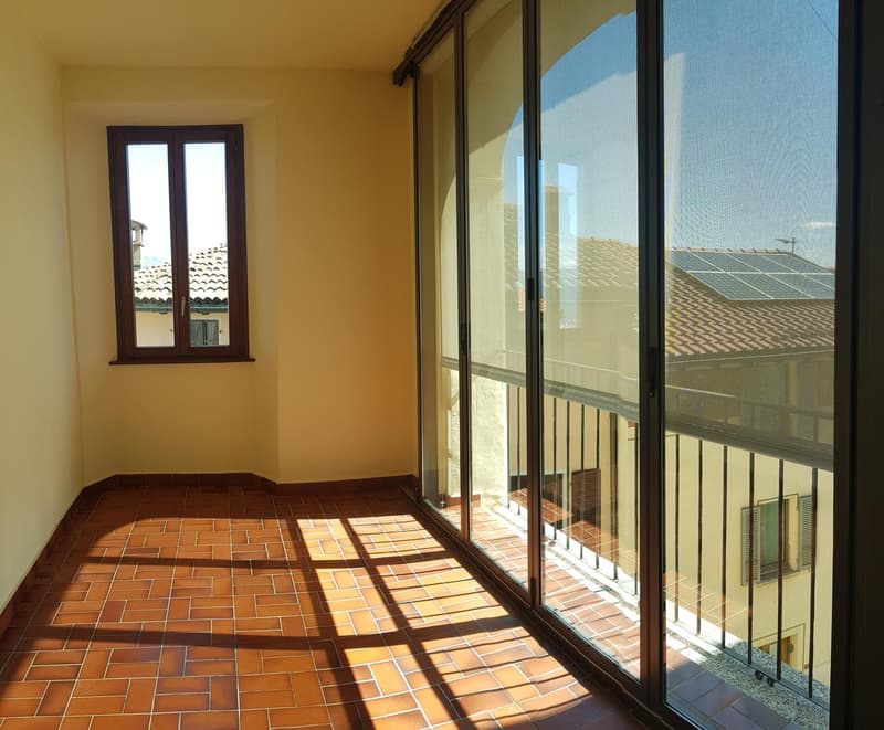Spacious room with large windows and sliding glass doors leading to a balcony or terrace. The floor is covered in terracotta tiles, creating a warm and rustic feel. The room has a simple, minimalist design with neutral-colored walls.