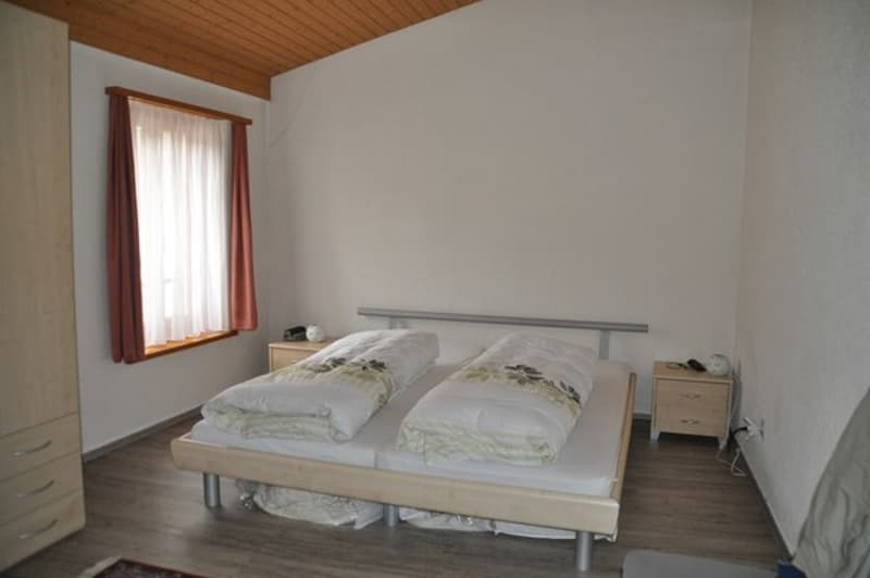 2 beds, white sheets, two nightstands, one has a phone, wooden floors, window with white curtains, wooden headboard, wooden wardrobe