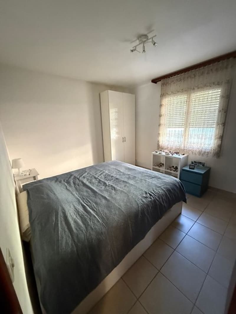 Bedroom with bed, white wardrobe, nightstand with lamp, blue chest of drawers, tiled floor, window with curtain, white ceiling with lamps