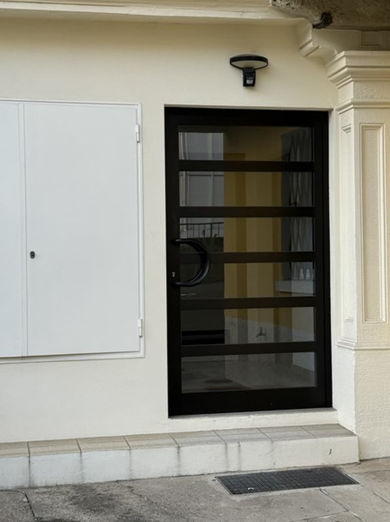 Front entrance to building, black door with glass panels, white door to the left, light above the door
