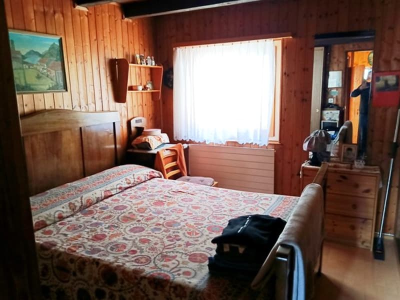 wooden walls, bed, wooden headboard, pillow, blanket, radiator, window with white curtains, picture frame, shelves with items, mirror, wooden chair, cabinet with drawers, table, bag, radiator, broom