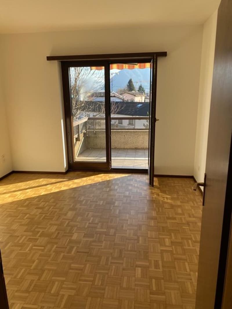 Spacious living room with hardwood parquet flooring, large sliding glass doors leading to a balcony or terrace, and a view of the outdoor scenery through the windows.