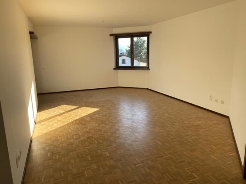 Spacious living room with hardwood flooring, white walls, and a large window providing natural light.