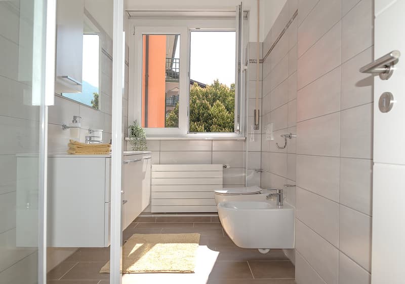 modern bathroom with white tiles, white vanity, white toilet, white sink, window, rug, towels