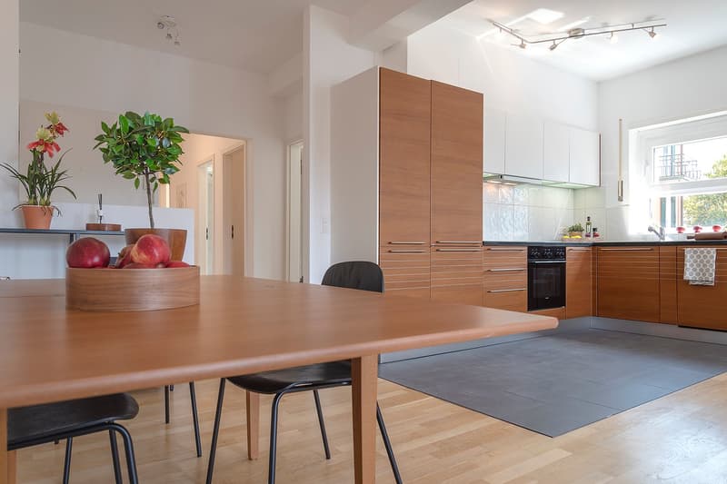 modern kitchen with wooden cabinets, white walls, wooden dining table, black chairs, potted plants, bowl of apples, black oven, sink, window