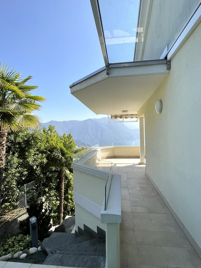 A balcony with a glass railing and a tiled floor, offering a view of the mountains and a water body. The balcony has a handrail, a light fixture, and a staircase leading to the ground. There are palm trees and other vegetation in the area.