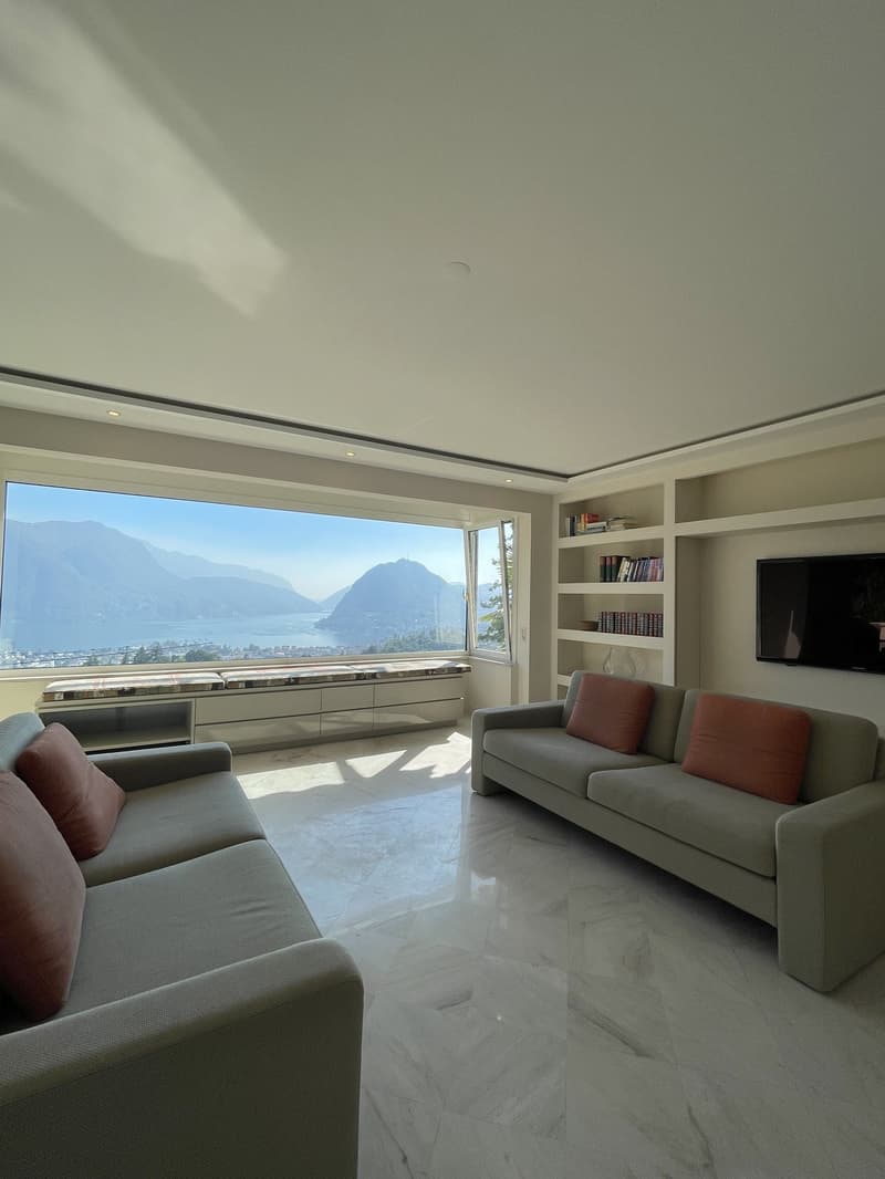 Spacious living room with large windows, two couches, and a window seat. The room features a stunning view of the mountains and a lake.