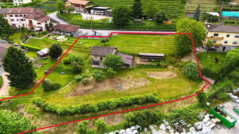 Empty plot of land with house in the middle, red line indicating boundary, surrounding are houses, cars parked, street light, trees, vineyard