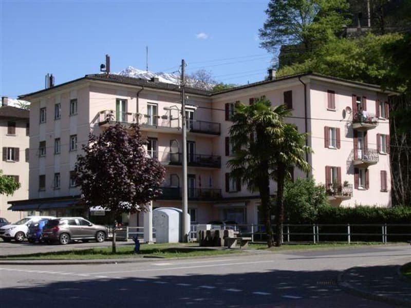 A multi-story building with balconies, multiple windows, and a flat roof. There are cars parked in front of the building, and a bench is placed in the grassy area. The building is surrounded by trees and bushes, and there is a mountain in the distance.