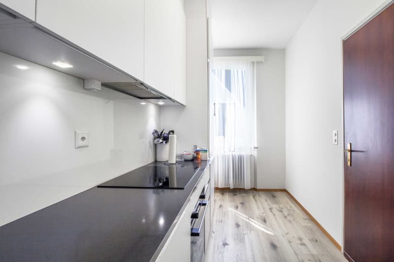 modern kitchen with white cabinets, black countertops, induction stove, white walls, wooden floor, white curtains, power outlet, and a window with a white curtain
