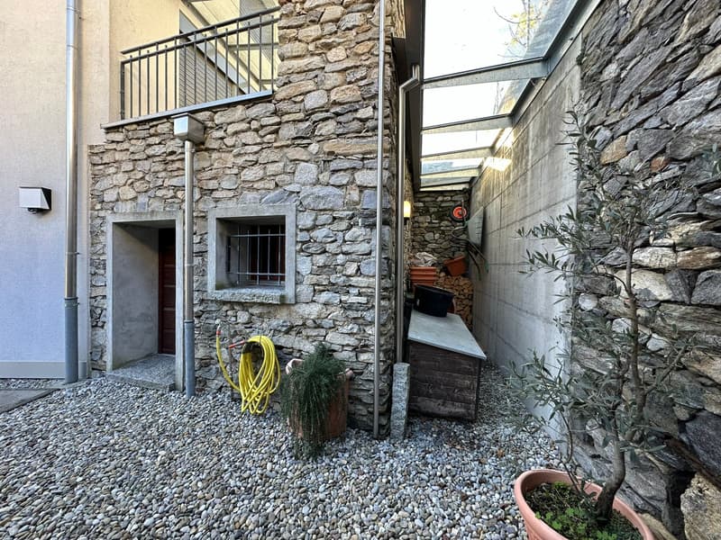 Stone building, raised ground floor, balcony with metal railing, entrance door, potted plants