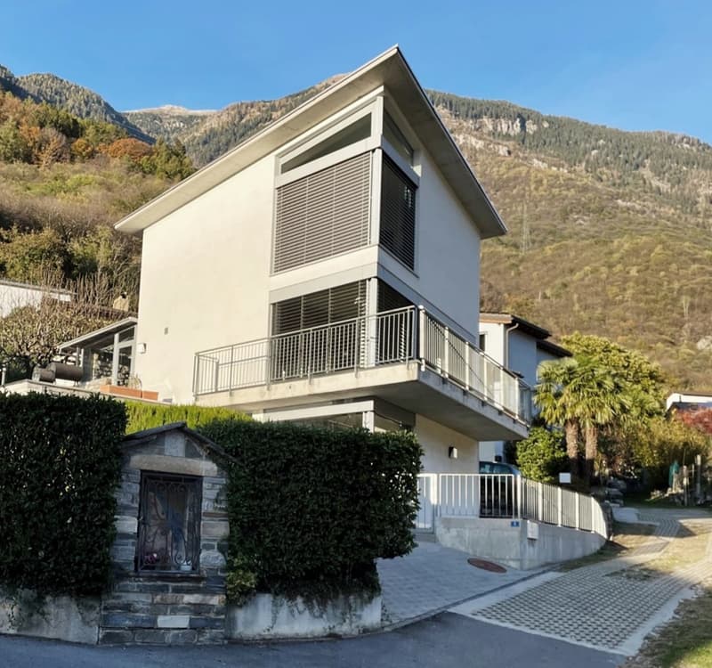 Modern 2-story house, white facade, balcony with metal railing, driveway with concrete curb, lush greenery, hilly background, clear sky