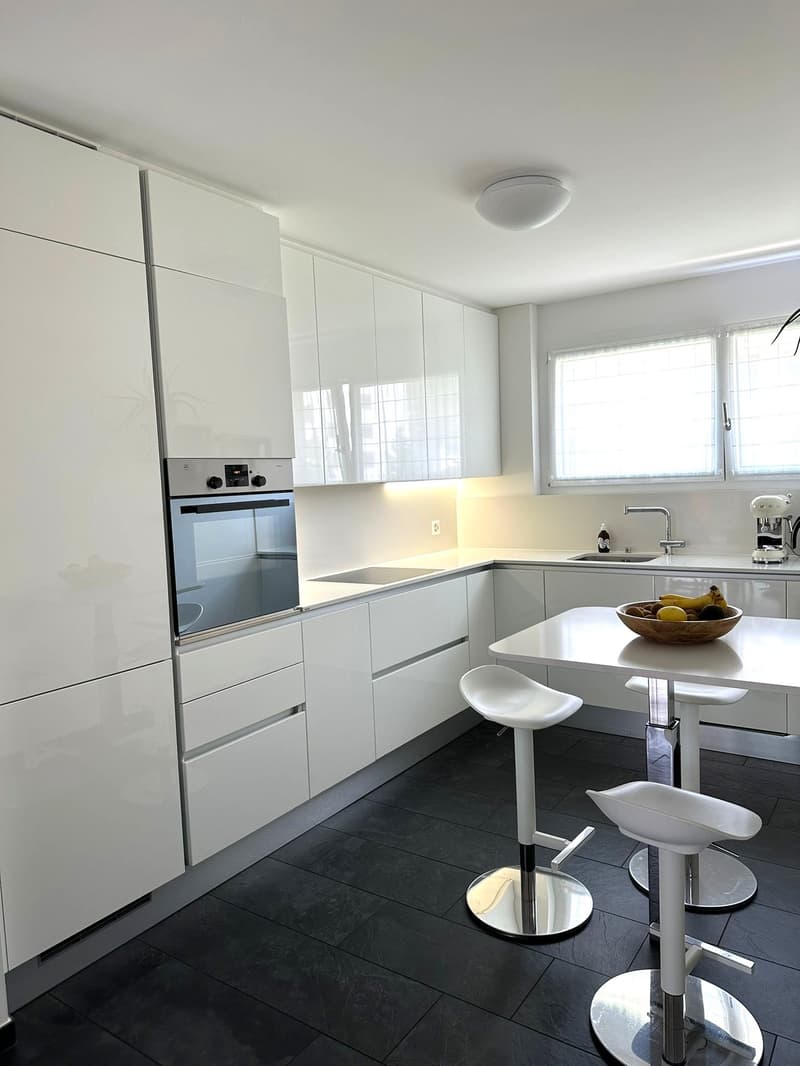 Modern, minimalist kitchen with white cabinets, stainless steel appliances including a dishwasher, white countertops, and dark tile flooring. The kitchen has a breakfast bar with white stools and a window providing natural light.