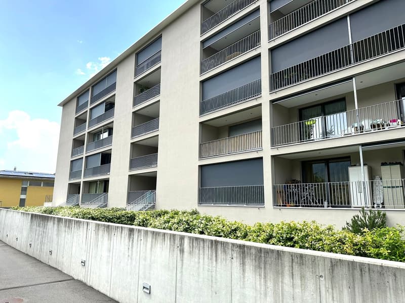 Multi-story apartment building with gray exterior, balconies on each floor, and landscaping around the base of the building.
