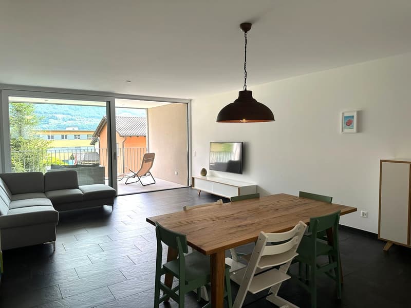 Spacious open-plan living area with a large sofa, wooden dining table and chairs, a TV stand, and a sliding glass door leading to a balcony or terrace with a scenic view of the surrounding landscape.