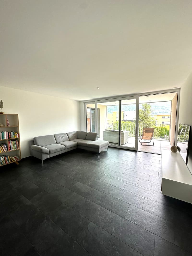 Spacious living room with large windows, grey sectional sofa, bookshelf, and sliding glass doors leading to a balcony or terrace.