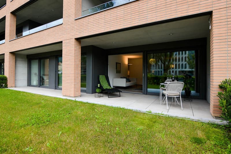 A brick building with glass doors leading to a patio, a black lounge chair, a small table, and a white chair, and a couch is inside. There is a grassy area and shrubs on the side.