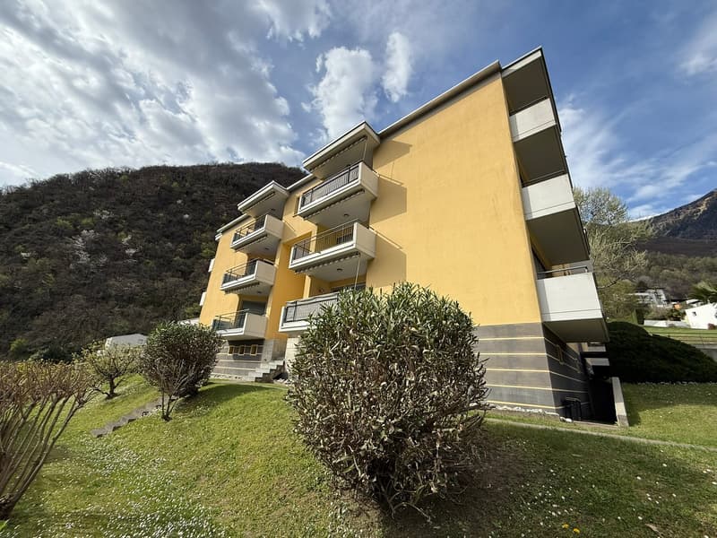 Yellow building with balconies on each floor, stairs leading up to building, small plants and bushes in front