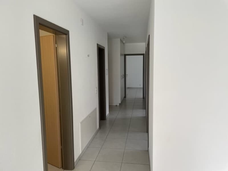 The image shows a long, narrow hallway with white walls and gray tile flooring. There are several doors visible, including a wooden door and a closed door. The hallway appears to be well-lit and clean.