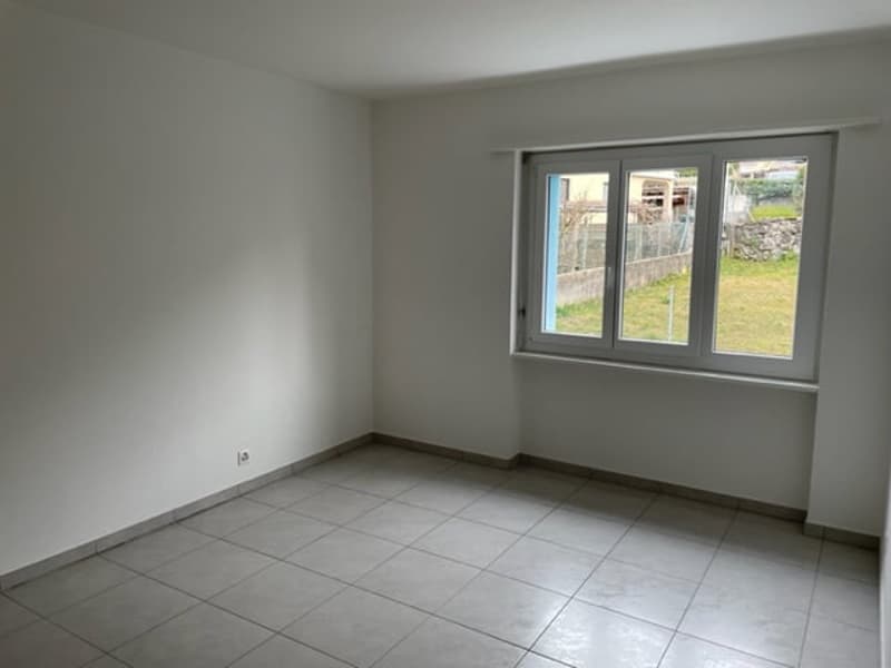 The image shows an empty room with white walls and a tiled floor. There is a large window that provides natural light and a view of the outdoor area.
