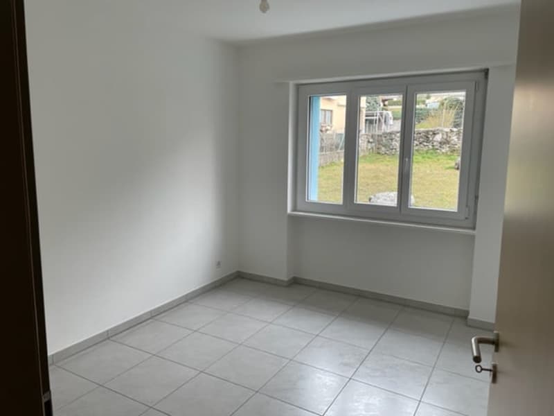This image shows a room with white walls, a tiled floor, and a large window overlooking a garden or outdoor area. The room appears to be empty, with no furniture or decor visible.