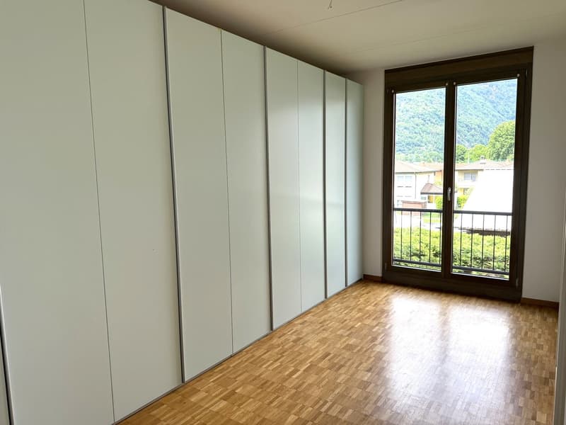 Spacious bedroom with large built-in wardrobes along one wall, hardwood flooring, and a large window overlooking the surrounding greenery.